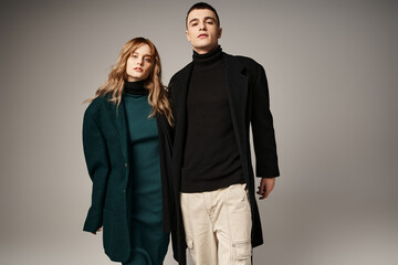appealing trendy boyfriend and girlfriend in coats posing and looking at camera on gray backdrop