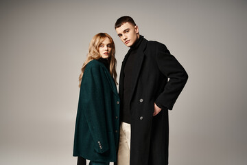 fashionable couple in stylish coats posing together on gray backdrop and looking at camera