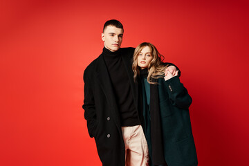 elegant woman in coat looking at camera and posing lovingly with her boyfriend on red backdrop