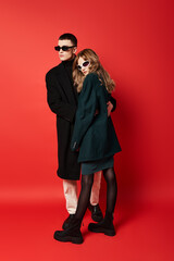 sophisticated young couple in stylish coats with sunglasses posing together on red backdrop