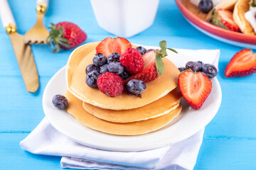 Stack of traditional american pancakes with summer berries. Breakfast sweet pancakes with honey, strawberry and blueberry, on morning sun lighted blue wooden background copy space