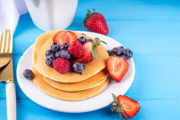 Stack of traditional american pancakes with summer berries. Breakfast sweet pancakes with honey, strawberry and blueberry, on morning sun lighted blue wooden background copy space