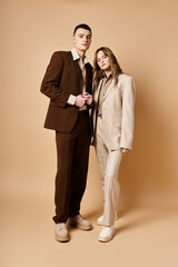 alluring fashionable couple in stylish suits posing on pastel backdrop and looking at camera