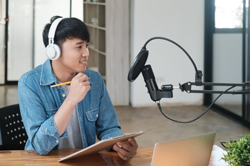A man wearing headphones is sitting at a desk with a laptop and a notebook. He is smiling and he is...