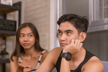 Eye-rolling young Asian man shows contempt while ignoring his girlfriend's concerns in a living room setting.