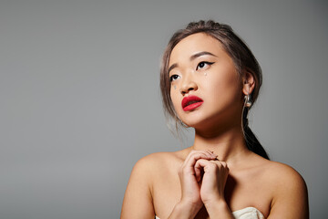attractive asian woman with red lips folding hands and looking to up against grey background
