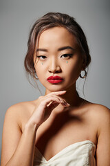 portrait of confident asian woman with elegant makeup touching to chin with hand on grey background