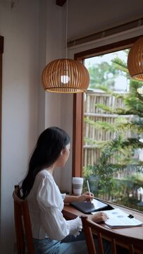 An Asian female entrepreneur working at a nearby café, diligently taking notes on her tasks using a tablet, demonstrating focused remote work