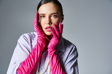 Dreamy woman in violet shirt and pearls, pink trendy gloves, touching her lips and standing on grey