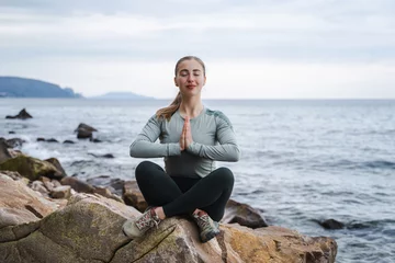 Zelfklevend Fotobehang Young calm smiling woman with closed eyes praying in lotus pose at the sea, girl in sport clothes relaxing and meditation outdoors, try to calm down, female keeping palms together, please God bless me © Соня Монштейн