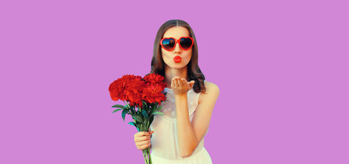 Beautiful young woman with bouquet of red rose flowers and blowing kiss on purple studio background