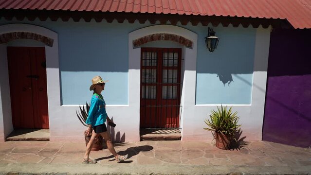Mature woman tourist wearing straw hat and ethnic, traditional clothes walking in front of blue home in mining town of El Triunfo, Mexico.