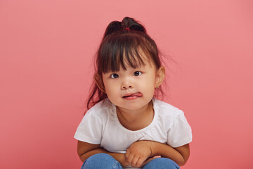 Funny little girl is squatting with her tongue sticking out on a pink isolated background. Cute...