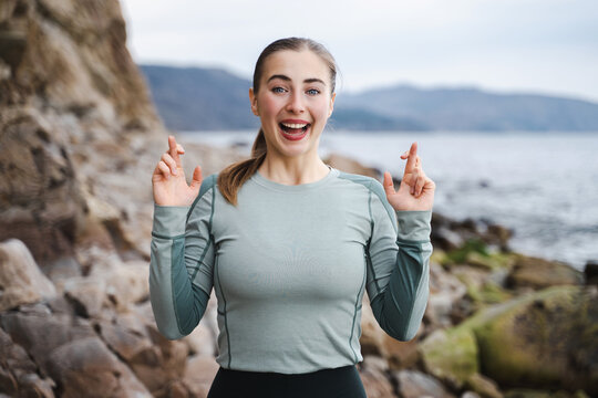 Young positive smiling woman looks at camera and keeps fingers crossed believes in good luck, sporty girl gesturing finger crossed with hopes to win, superstitious concept