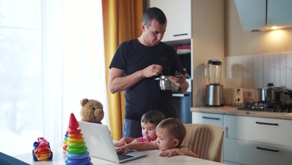 father cooking in the kitchen remote work. baby twins playing with laptop in the kitchen. father working at home in the kitchen stirring food in the pan. remote work business lifestyle concept