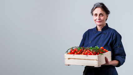 An older chef in a blue uniform holding a crate of tomatoes tenderly on a white backdrop