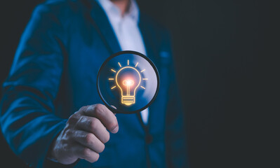 Businessman holding a magnifying glass that illuminates a light bulb icon, representing insight, innovation, and the search for bright ideas in the corporate world. Business solution idea concept.