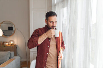 A contented young adult man having a cup of while relaxing at home and looking through a window.