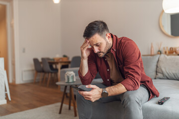 Fototapety  An anxious adult man reading bad news on his phone while sitting on a couch