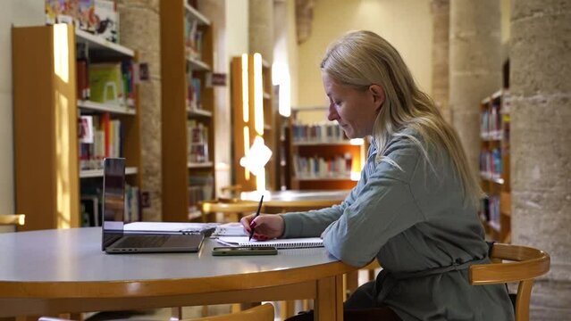 Adult female student prepare material essay for educational course for college or university in library. Middle aged woman using laptop, researching for materials, essay, test or exam training.