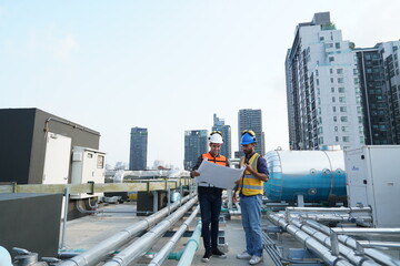 Foreman engineer and contractor wearing reflective jacket, engineering helmet, holding blueprints and walkie-talkie, inspects construction work and plumbing utilities on building rooftops.