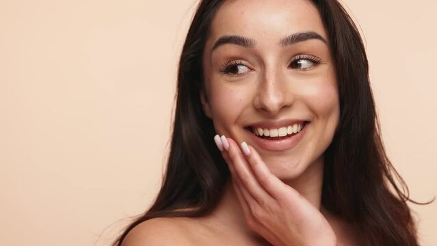 Close-up of a joyful young woman with clean skin gently touching her face, embodying a beauty and skincare concept with a neutral studio background.