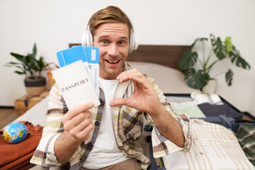 Portrait of handsome smiling man in headphones, sits with opened suitcase on bed, shows passport with two plane tickets, recommends travel agency, goes on holiday, excited about vacation - obrazy, fototapety, plakaty