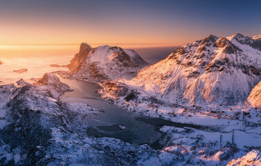 Aerial view of snowy mountains, blue sea and orange sky at sunset