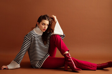 Fashionable confident woman wearing stylish striped woolen sweater, red jeans, patent leather  ankle boots, posing on brown background. Studio fashion portrait. Copy, empty, blank space for text
- 767177689