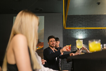 Man in black shirt raises glass of cocktail while making toast pointing at blondes at bar counter....