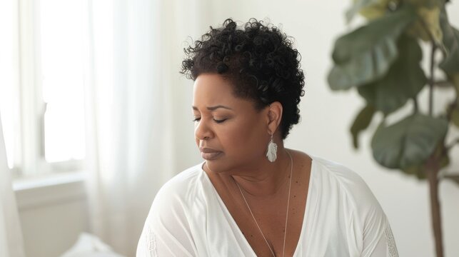 A woman with curly hair wearing a white blouse and earrings looking down with a thoughtful expression set against a bright blurred indoor background with a plant.