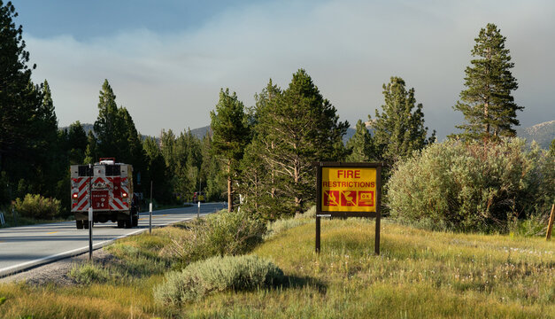 Fire restrictions sign in Sierra Nevada forest with fire truck passing by and smoky skies from nearby fires actively burning.  The image captures the urgency of wildfire prevention.