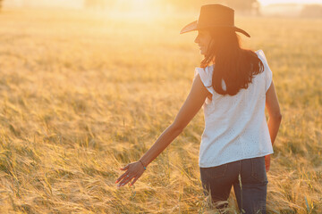 Woman farmer in cowboy hat walking with hands on ears at agricultural barley field on sunset. - 767209851