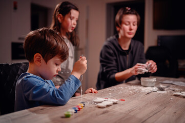 A young boy with a paintbrush, flanked by his family, deeply focused on painting, portraying a...