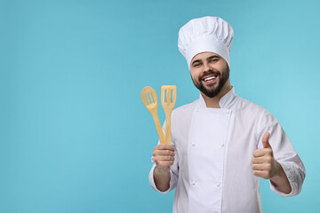 Happy young chef in uniform holding wooden utensils and showing thumb up on light blue background. Space for text