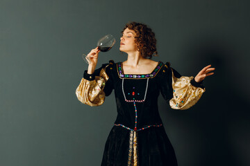 Young adult woman dressed in a medieval dress holding a glass of wine and drinking a red wine - 767210894