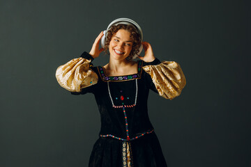 Portrait of a smiling young aristocratic woman with headphones and touching head and dressed in a medieval dress listens music - 767211001
