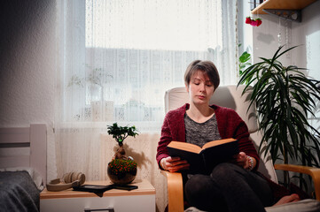 A serene woman deeply engrossed in reading a book, seated in an armchair, surrounded by lush houseplants in a warmly lit, peaceful home setting