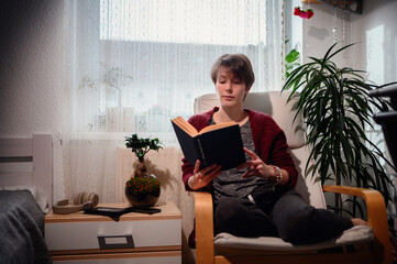 A serene woman deeply engrossed in reading a book, seated in an armchair, surrounded by lush...
