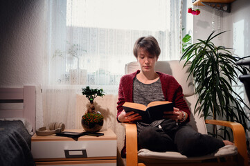 A serene woman deeply engrossed in reading a book, seated in an armchair, surrounded by lush...