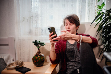 A young woman, dressed in casual home attire, focuses intently on her smartphone in a well-lit, cozy corner of a room, accompanied by a bonsai and modern headphones