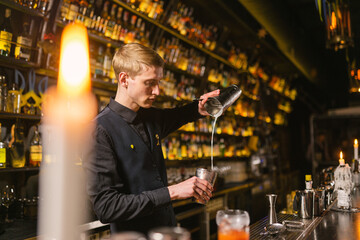 Professional barkeeper decants alcoholic drink into shaker. Young barman works night shift in Ukrainian bar with burning candle in foreground