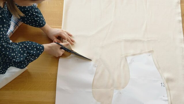 Close-up of a tailor's hands meticulously cutting beige fabric on a wooden surface using a paper pattern.
