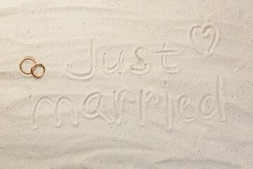 Honeymoon concept. Two golden rings and phrase Just married written on sand, top view