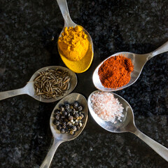 Five stainless steel teaspoons, filled with various types of spices, in a star shape on a dark...