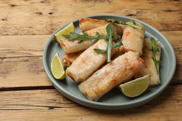 Plate with tasty fried spring rolls, arugula and lime on wooden table, closeup