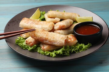 Plate with tasty fried spring rolls, lettuce, lime and sauce on light blue wooden table, closeup
