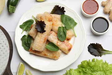 Plate with tasty fried spring rolls, spinach, sauces and other products on white tiled table, flat lay