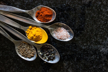 Five stainless steel teaspoons, filled with various types of spices, on a dark colored kitchen...