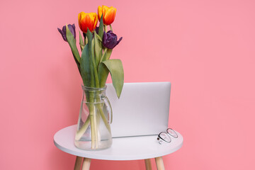 Bouquet of tulips in a glass vase on a white round table with a laptop in front of a pink background. - obrazy, fototapety, plakaty